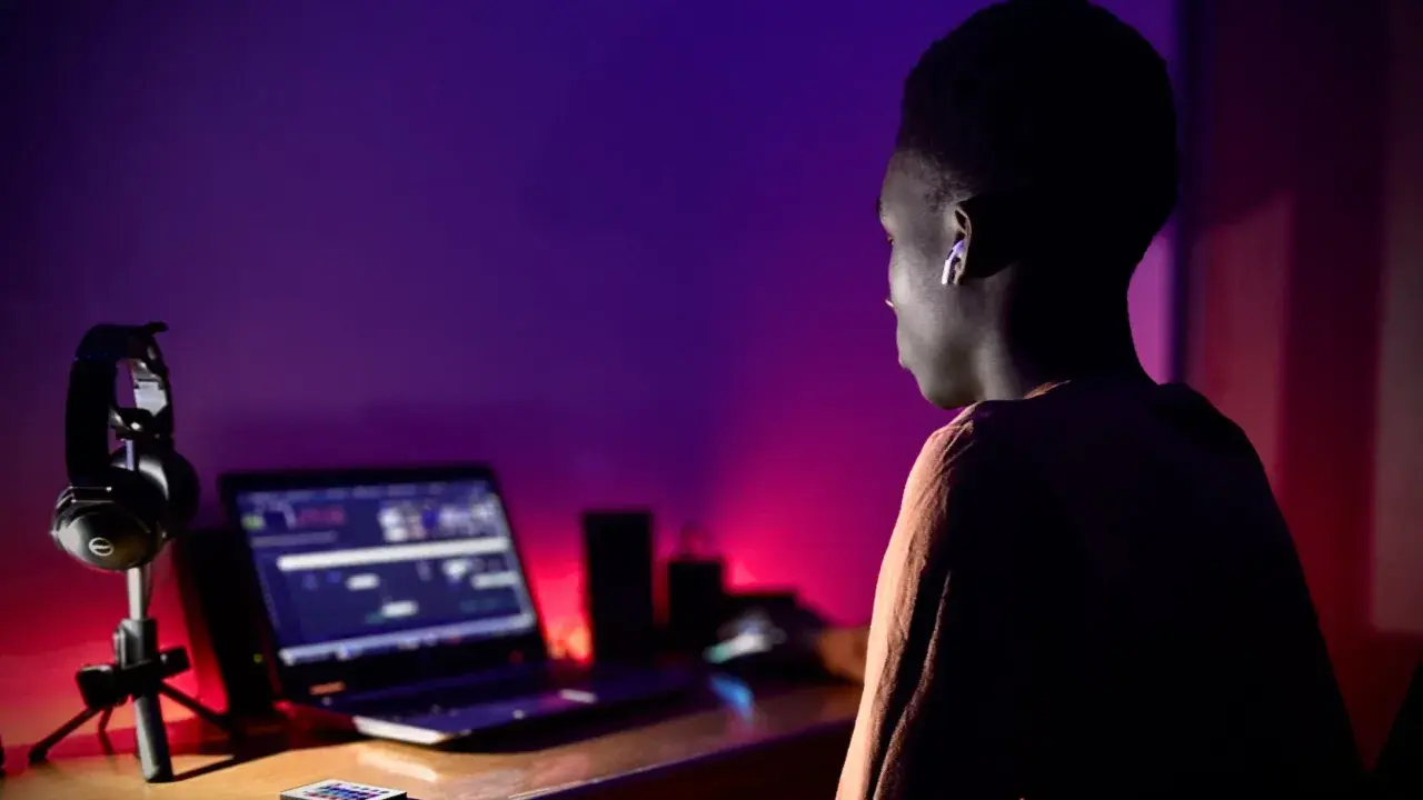 Un jeune homme, casque sur les oreilles, est assis devant un ordinateur portable, prêt à créer un son unique dans une ambiance tamisée aux lumières violettes et rouges.