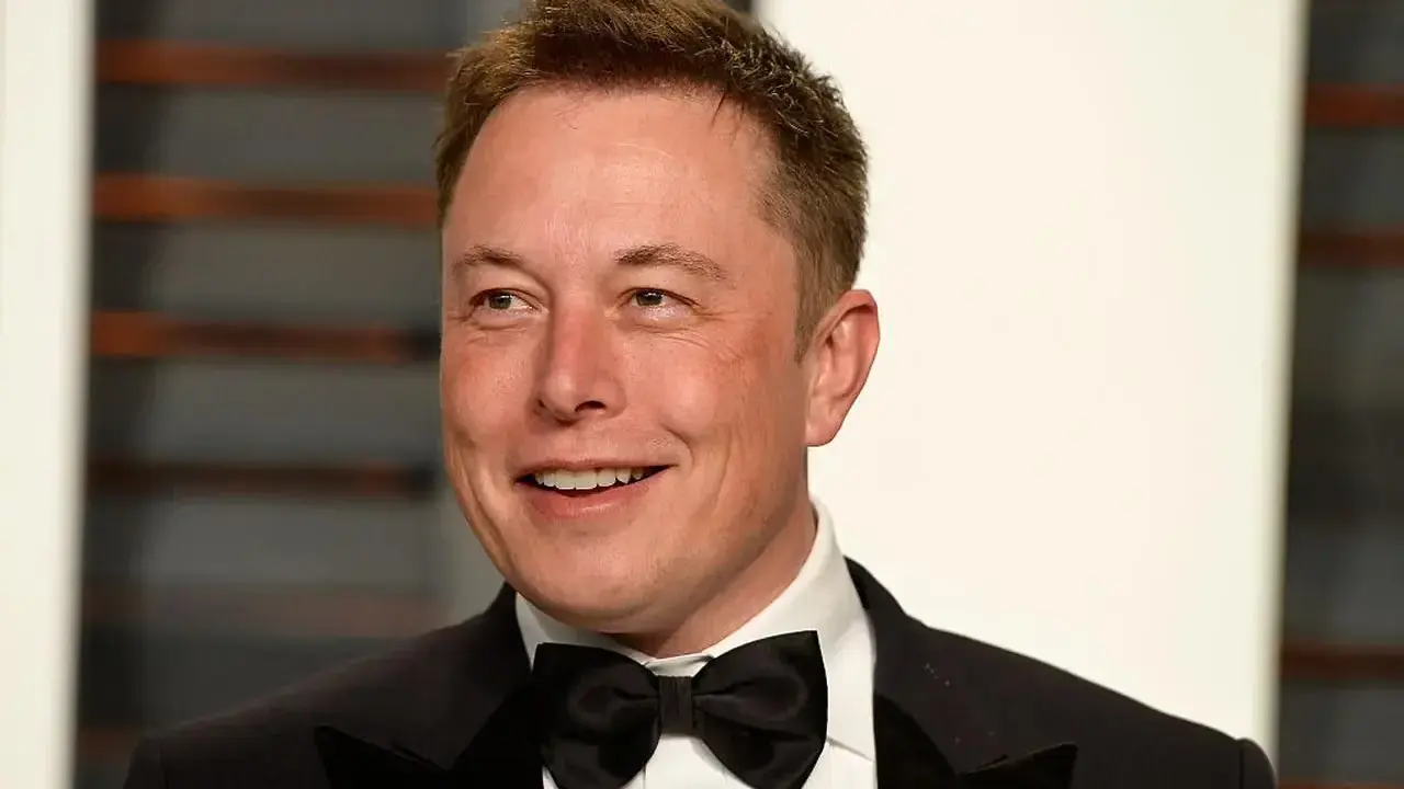 Uśmiechnięty Elon Musk w smokingu z muszką. Wiek nie jest widoczny, ale jego twarz wyraża pewność siebie.