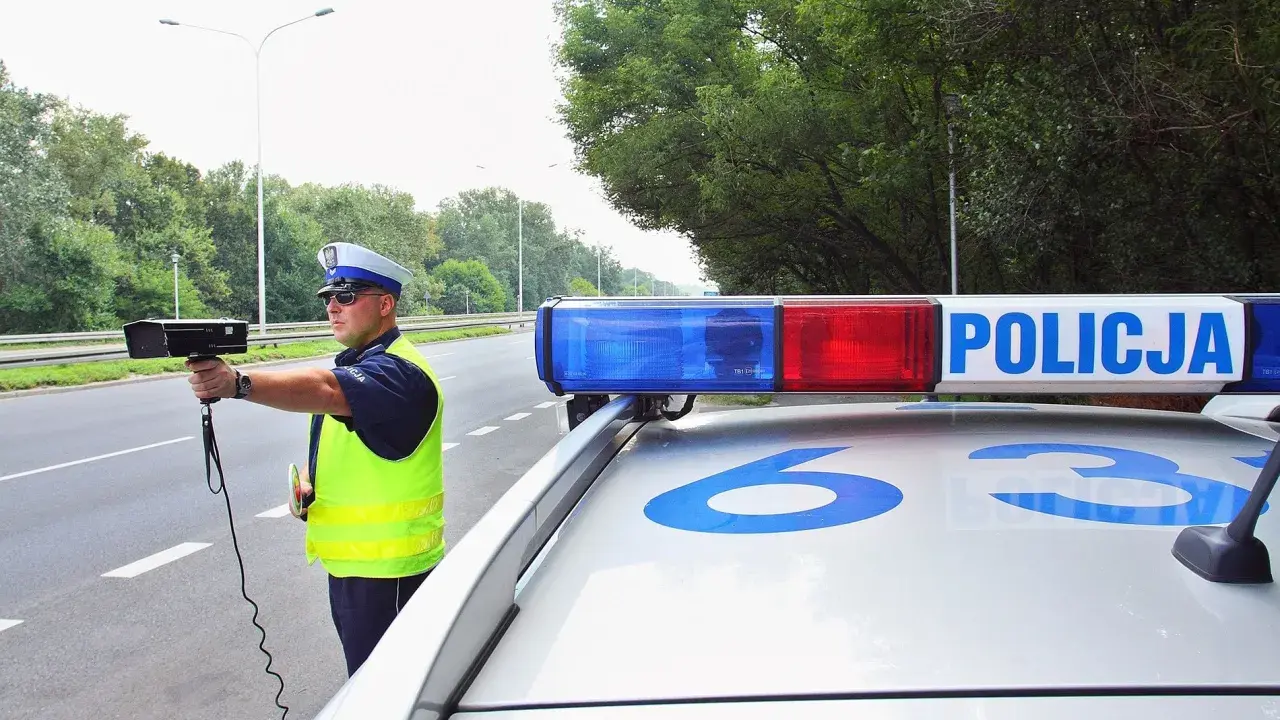 Policjant z radarem na drodze, obok radiowozu z napisem POLICJA.