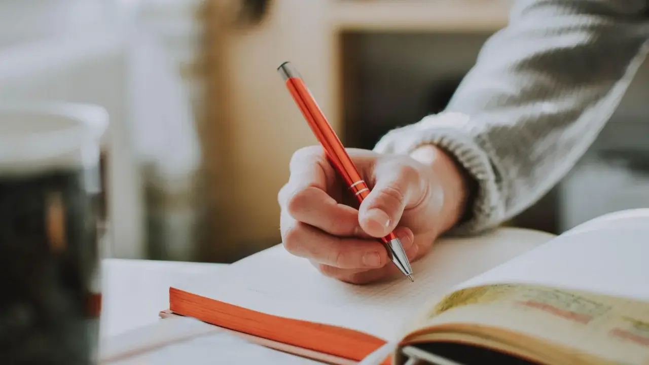 Main tenant une plume rouge, une main écrit dans un carnet. Un synonyme de participer pourrait être collaborer, comme dans cette scène d'étude.