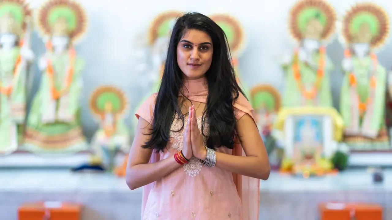 En ung kvinna i traditionella kläder med händerna i bön framför hinduiska statyer, en bild som väcker tankar om mantra hinduism.
