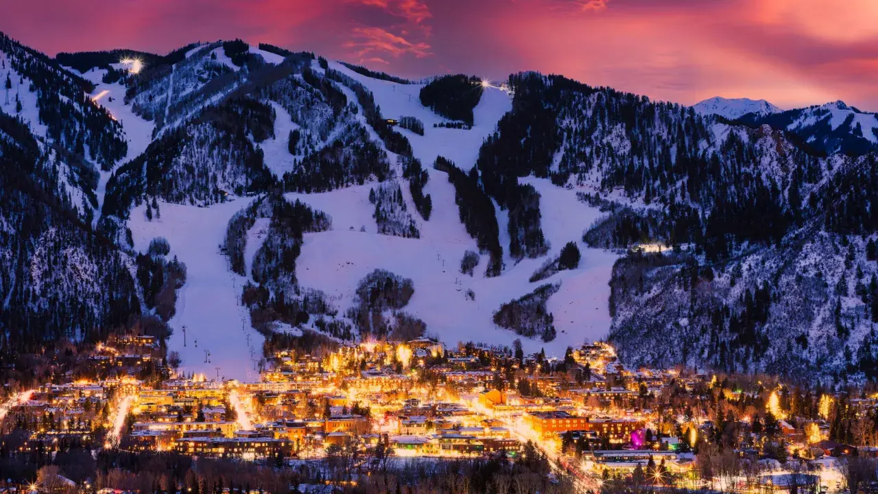 Glitzerndes Aspen, Colorado, im Abendrot. Schneebedeckte Berge mit Skipisten über einer Stadt, die in warmem Licht erstrahlt.