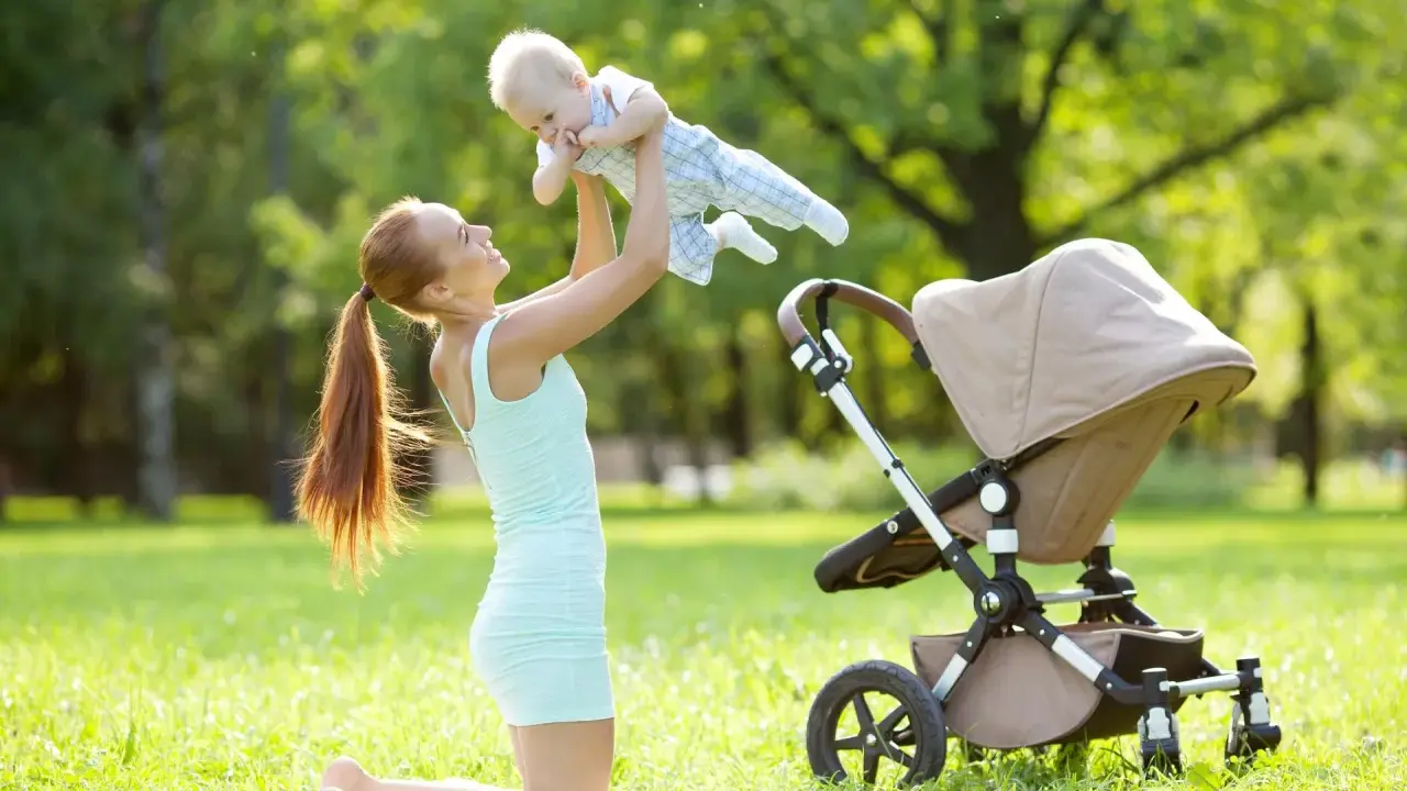 Uśmiechnięta mama podrzuca dziecko w powietrze na trawie obok wózka.