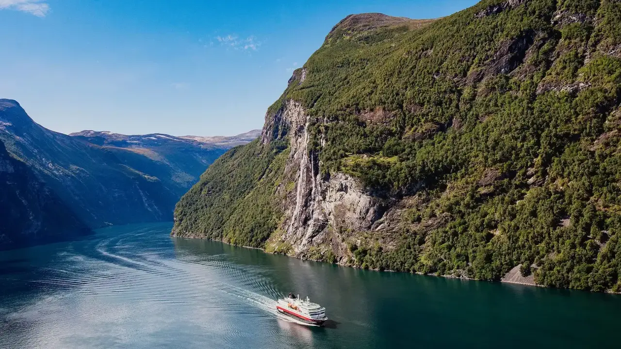 Ein Schiff fährt durch einen norwegischen Fjord im August. Das Wetter ist sonnig, die Berge sind grün und ein Wasserfall stürzt herab.