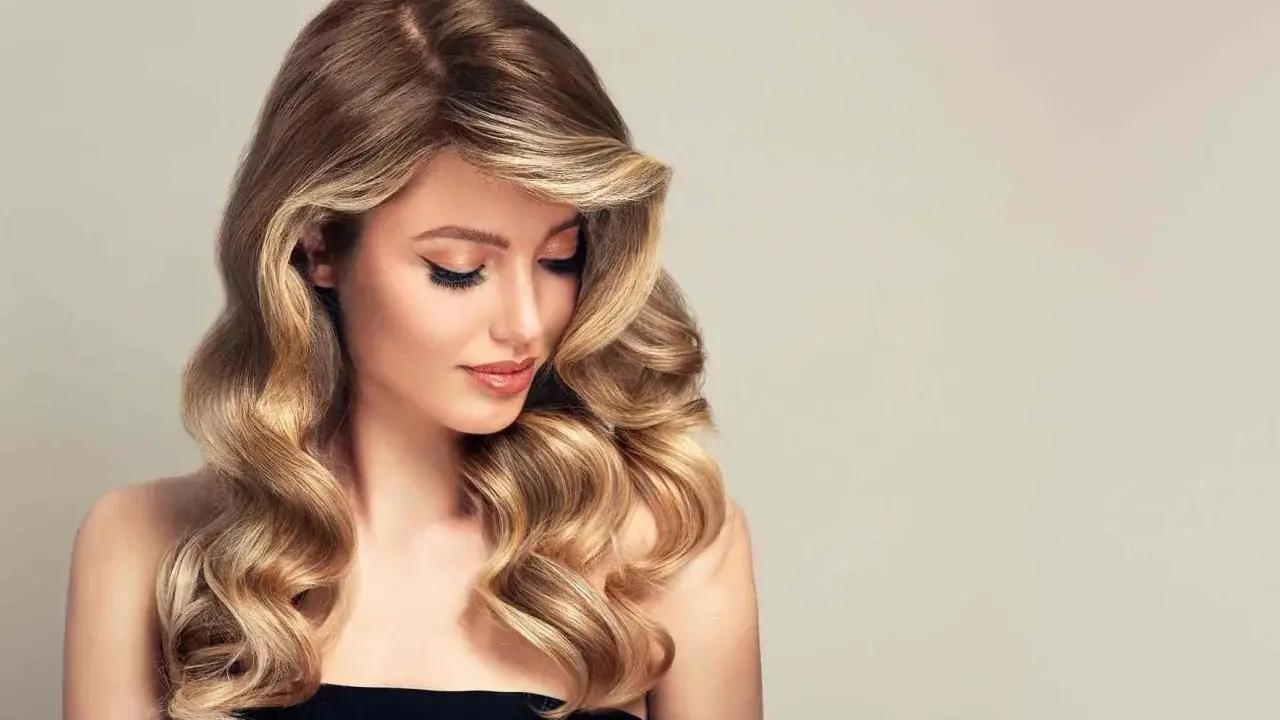 Kobieta z falowanymi, brązowo-blond włosami, delikatnym makijażem i spojrzeniem skierowanym w dół.