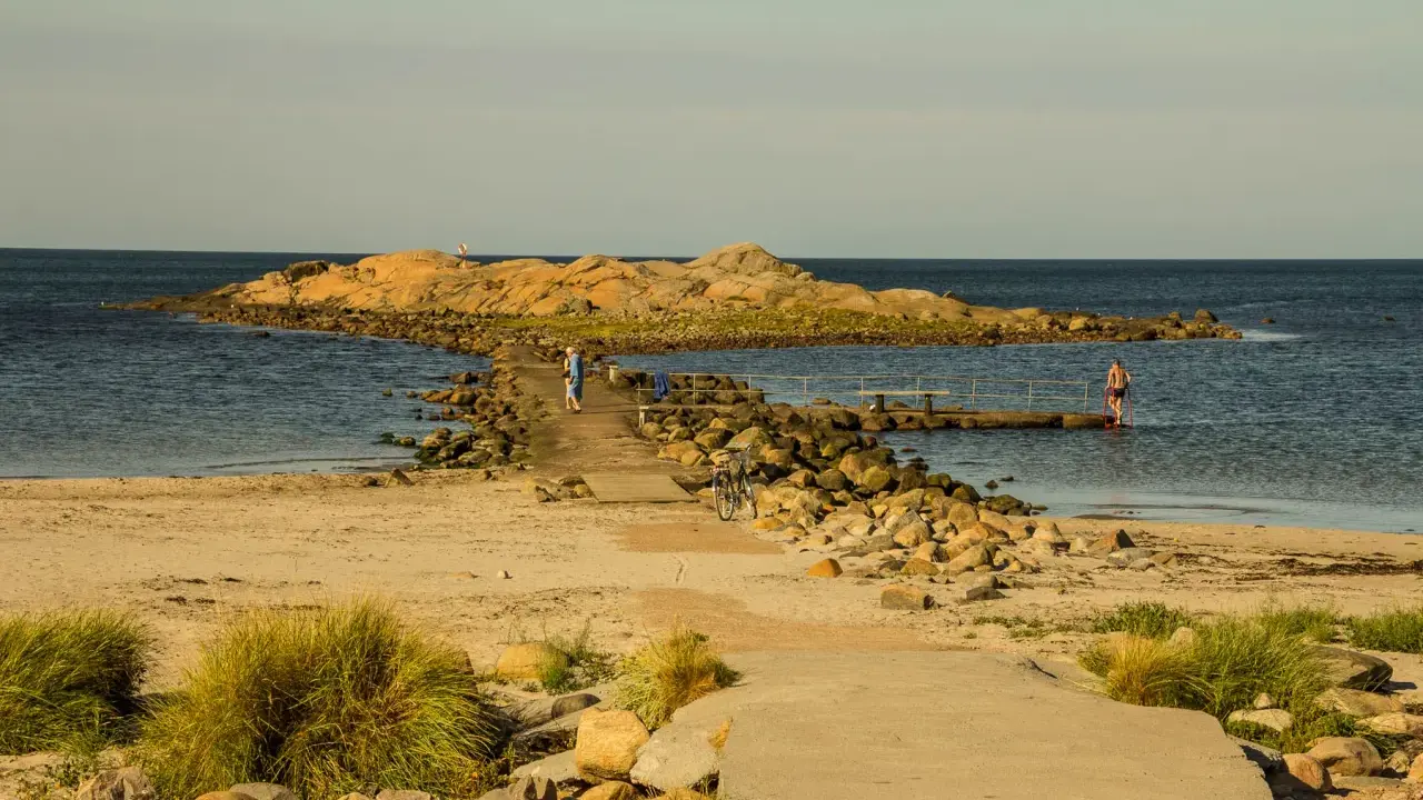 En stenpir leder ut till en klippö. En person går på piren, en annan badar. Perfekt för aktiviteter i Halmstad.