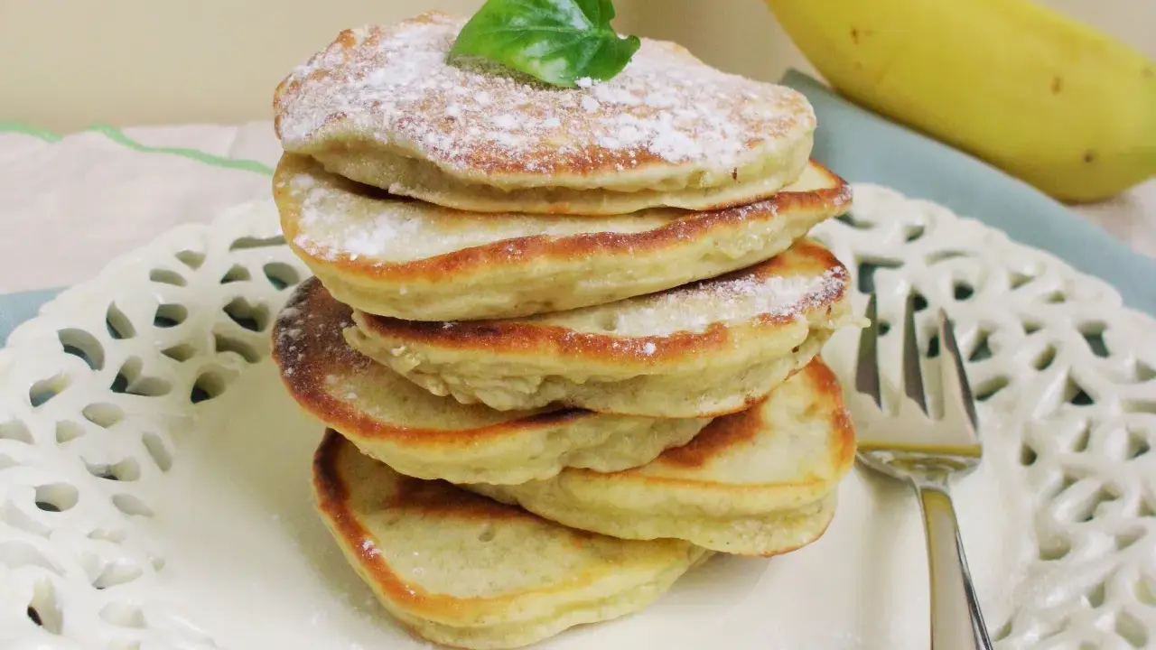 Stos puszystych, trzyskładnikowych placków bananowych, posypanych cukrem pudrem i udekorowanych listkiem, obok dojrzałego banana.