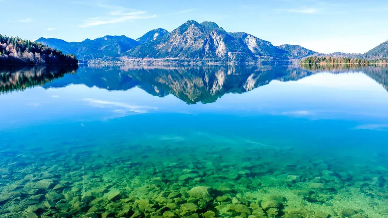 Kristallklarer Badesee in Bayern mit Felsen am Ufer und spiegelnden Bergen im Hintergrund.