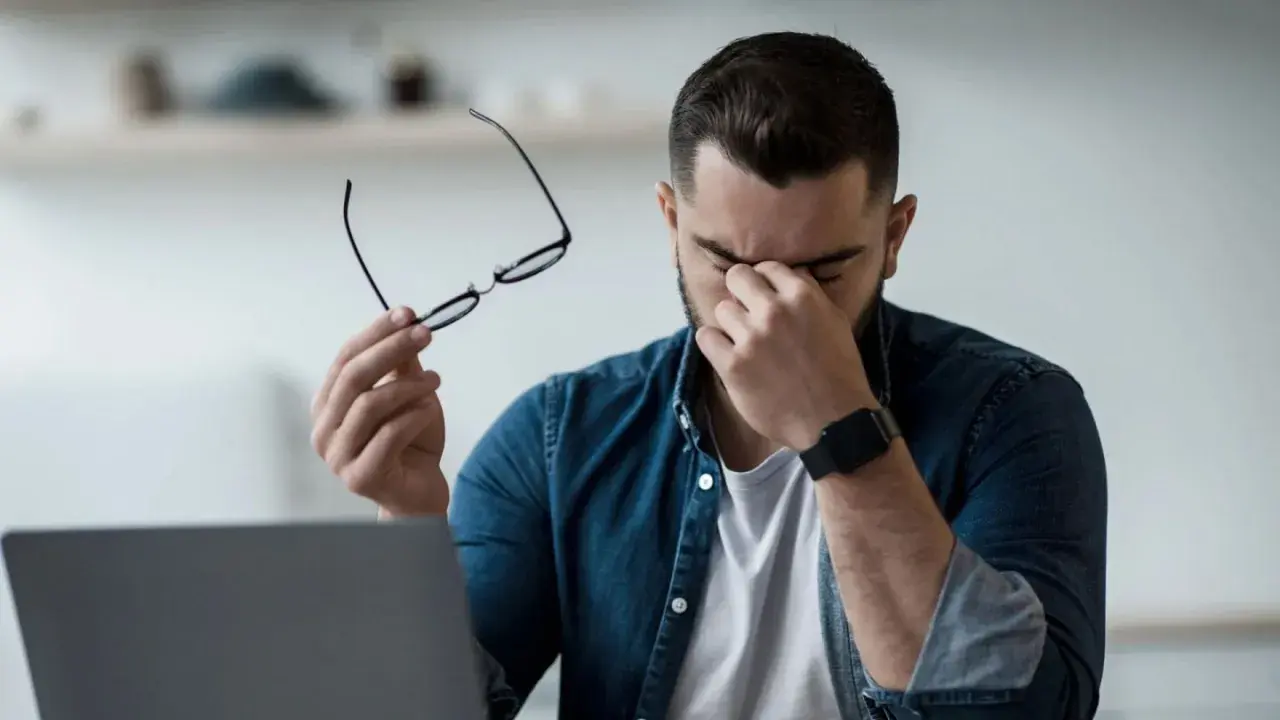 Homme fatigué, lunettes à la main, se frottant les yeux devant un ordinateur. Il est temps de penser à comment protéger ses yeux des écrans.