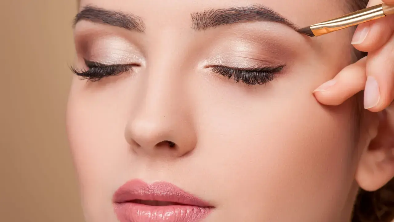 Une main applique un maquillage subtil pour parfaire la forme sourcil d'une femme aux yeux fermés.