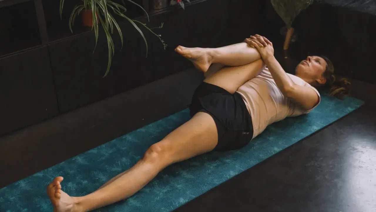 Femme effectuant un étirement psoas sur un tapis de yoga bleu. Elle est allongée sur le dos, une jambe pliée et ramenée vers la poitrine.