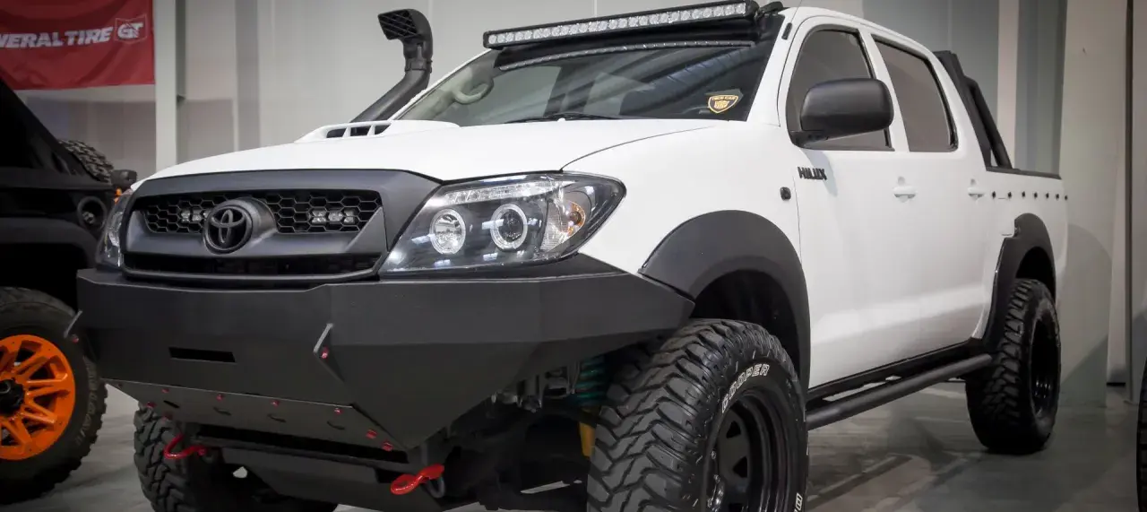 Biała Toyota Hilux z potężnym stalowym zderzakiem, wyciągarką i snorkel'em. Idealna do terenowych wypraw, gdzie przepisy nie ograniczają.