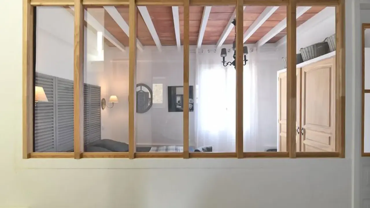 Une verrière en bois sépare deux pièces. On y voit un lit, des volets, une armoire et un lustre.