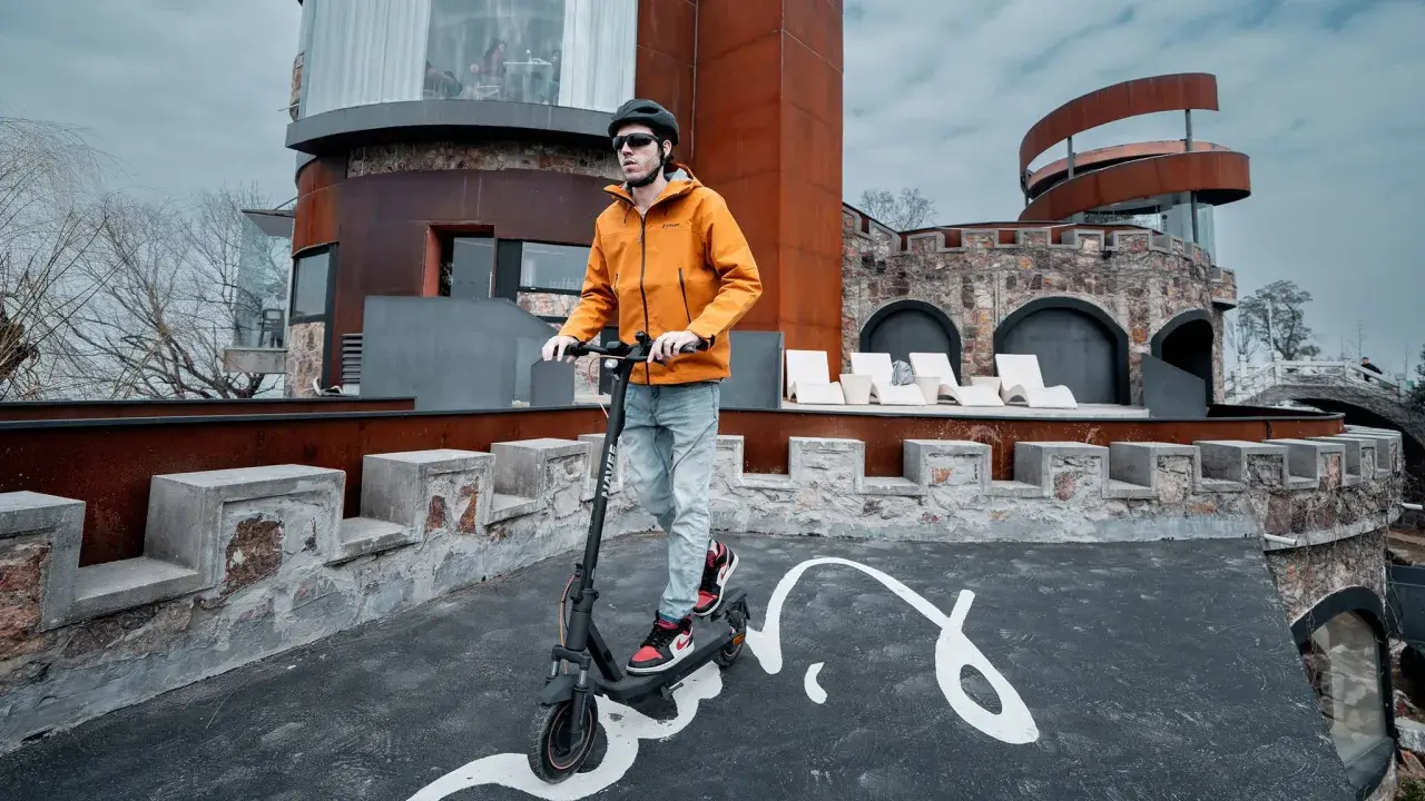 Un homme en casque et veste orange teste la vitesse de sa trottinette électrique devant un bâtiment de style médiéval moderne.