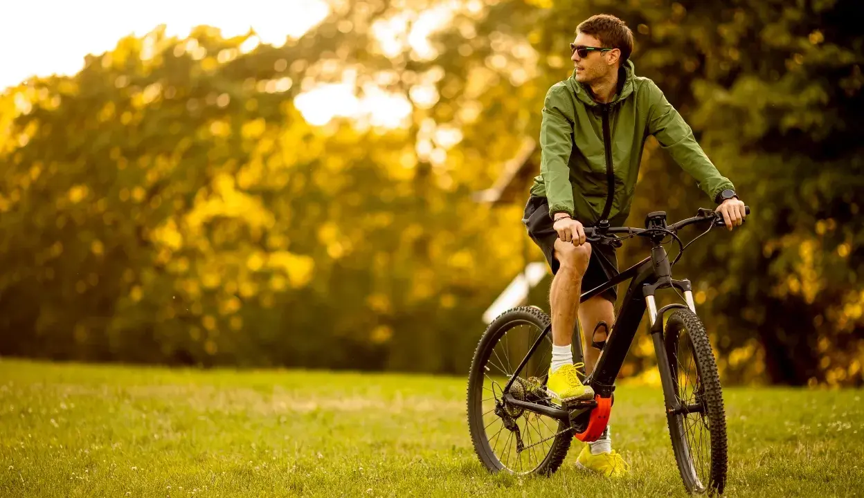 Mężczyzna w zielonej kurtce i okularach przeciwsłonecznych odpoczywa na trawie obok swojego roweru elektrycznego ebike.