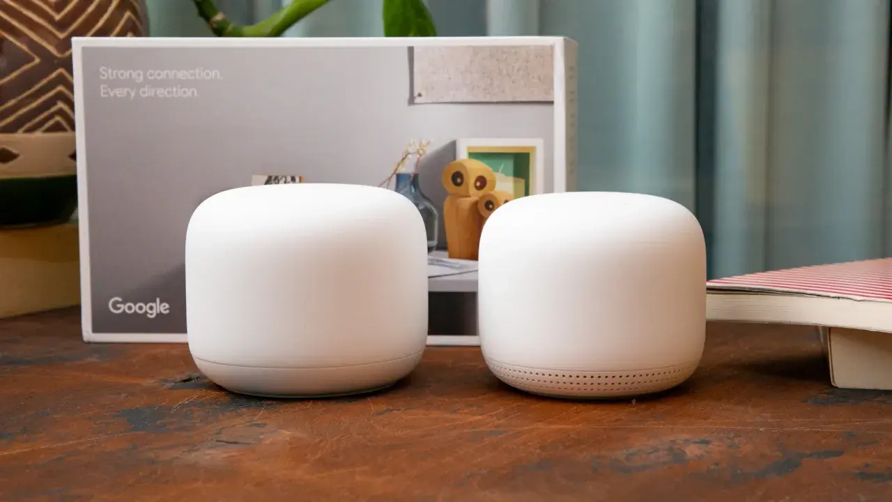 Två vita Google Nest Wifi-enheter står på ett träbord. En kartong med texten "Strong connection. Every direction." och Google-loggan syns i bakgrunden.