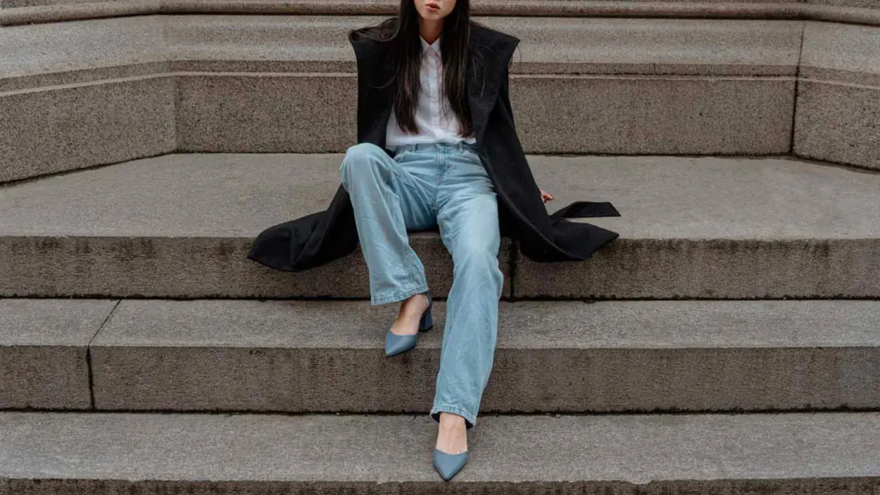 Donna con cappotto nero, camicia bianca e **pantaloni a sigaretta** chiari, indossa **tronchetti** blu.