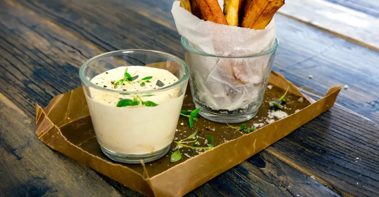 En krämig creme fraiche sås med örter serveras bredvid en glasbehållare med krispiga pommes frites på ett rustikt träbord.