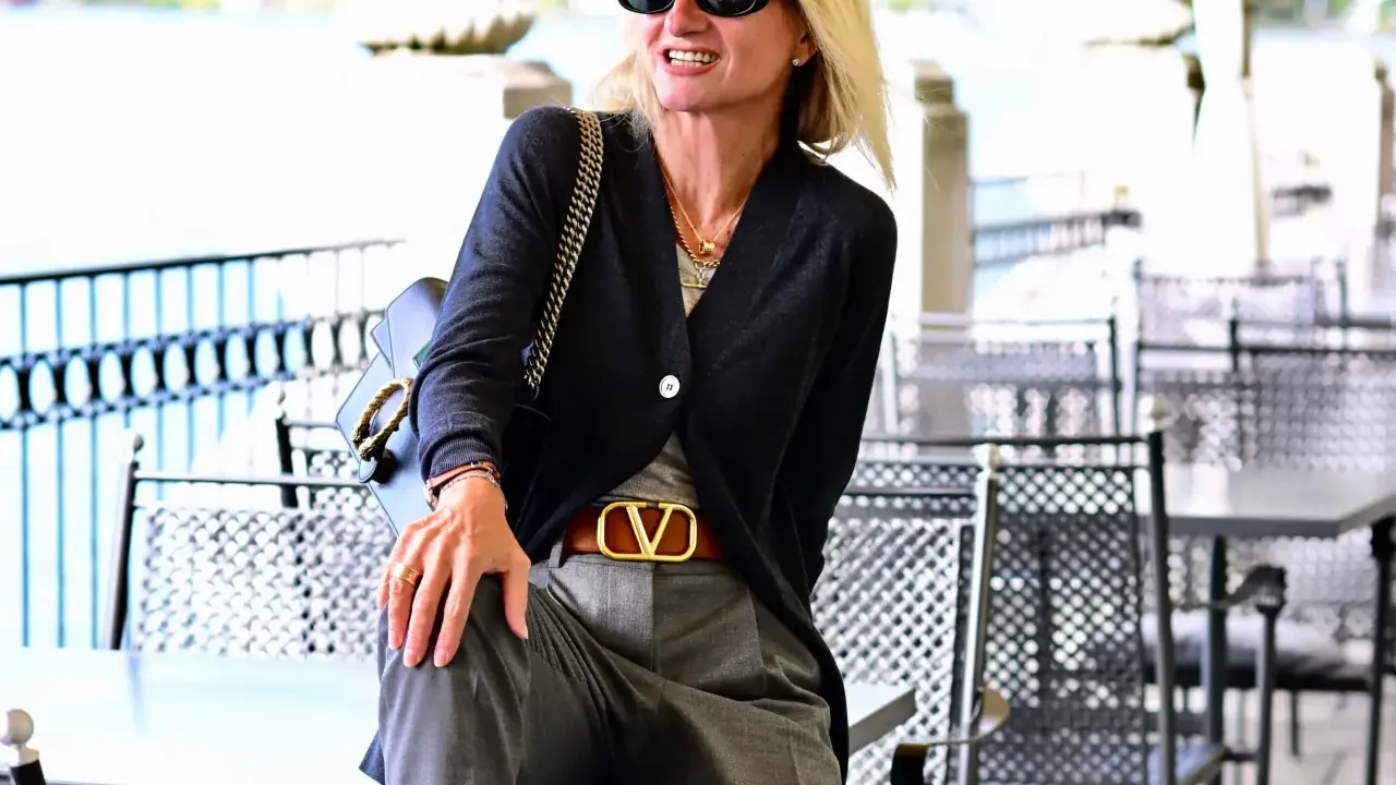 Donna con pantalone grigio elegante, abbinato a cardigan scuro e cintura con logo dorato. Un look chic per ogni occasione.