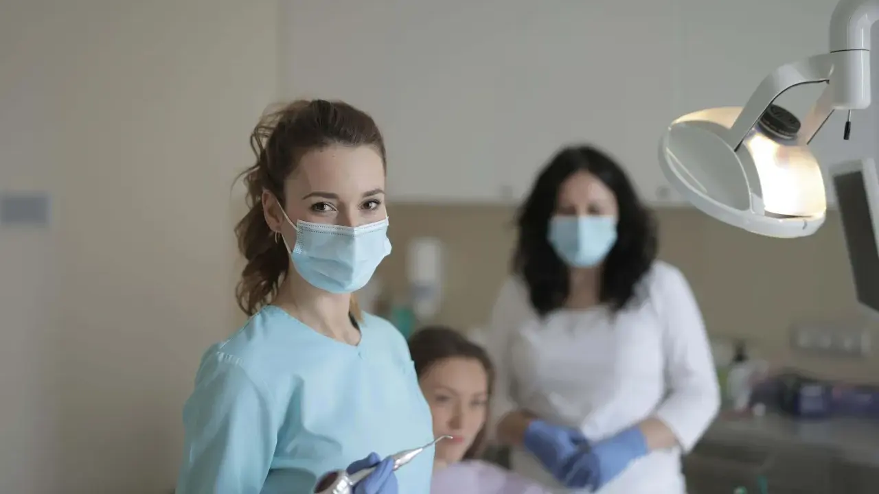 Uśmiechnięta studentka stomatologii w masce i rękawiczkach trzyma wiertło dentystyczne. W tle inne osoby w gabinecie.