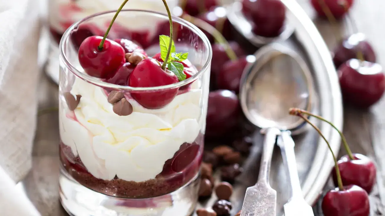 Delicioso postre con capas de crema batida, bizcocho de chocolate y cerezas frescas, coronado con más cerezas y trocitos de chocolate.