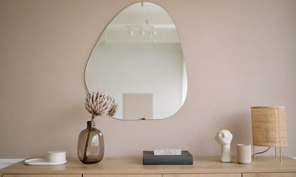Un miroir vitrail aux formes organiques reflète une pièce épurée. Une fleur séchée dans un vase en verre fumé et une lampe en rotin complètent la scène.