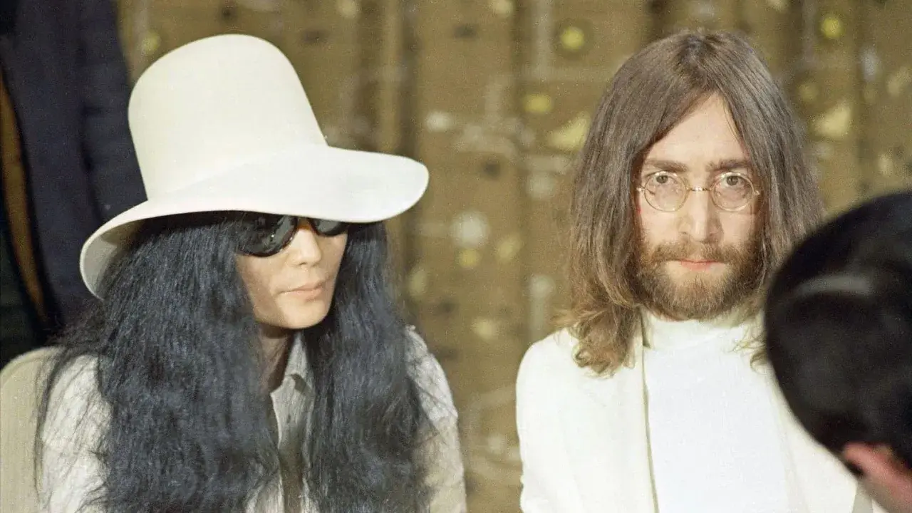 John Lennon och Yoko Ono, ikoniska konstnärer, poserar tillsammans. Yoko bär en stor vit hatt och solglasögon, medan John har sitt karaktäristiska långa hår och glasögon.