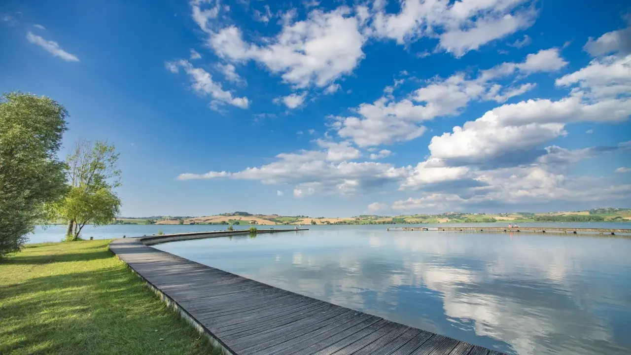 Passeggiata in legno sul **lago di Chiusi**, con cielo azzurro e nuvole bianche che si riflettono sull'acqua calma.