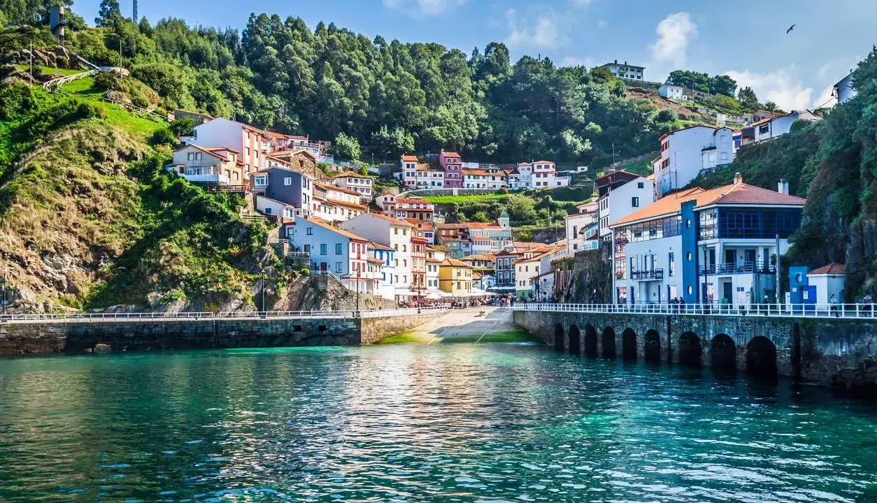 Un pintoresco pueblo costero con **5 villas** coloridas apiladas en una colina verde, frente a un mar turquesa.