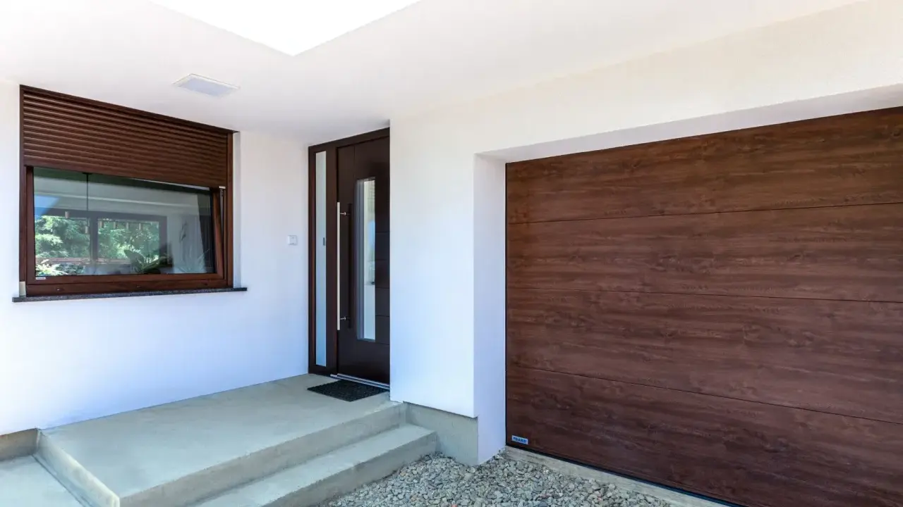Ingresso con porta scura e avvolgibili marroni, accanto a un'ampia porta garage in legno scuro.