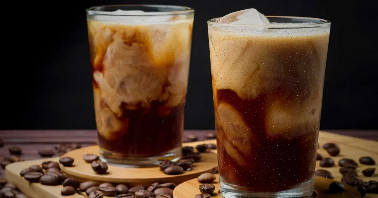 Två glas med iskaffe, perfekt för ett iskaffe recept. Krämig mjölk virvlar i mörkt kaffe med isbitar. Kaffebönor sprider sig runt glasen.