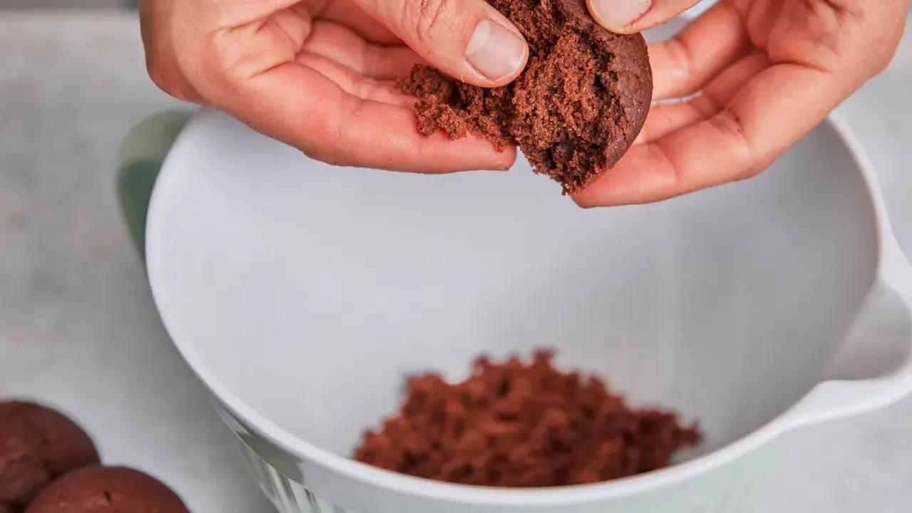 Hände zerbröseln einen Schokoladenkuchen für vegane Desserts. Ein Teil landet in einer weißen Schüssel.