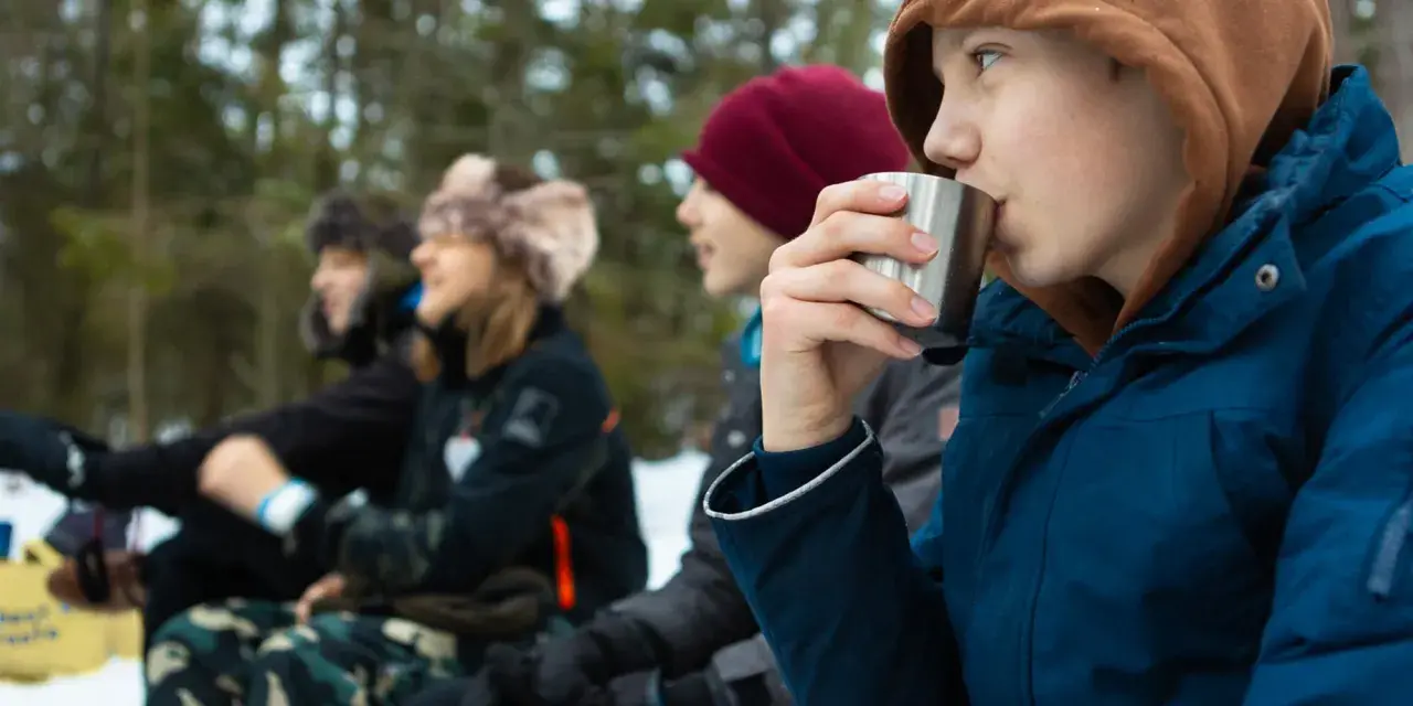 Barn njuter av fika under friluftsdag aktiviteter i snöig skog. En pojke dricker ur en metallmugg.