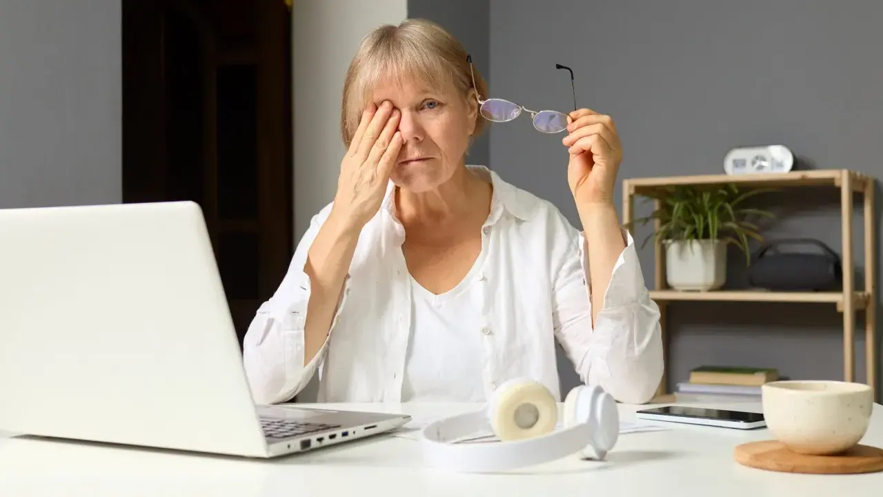 Zmęczona kobieta pociera oko, patrząc na ekran laptopa, który emituje niebieskie światło. Obok leżą okulary i słuchawki.