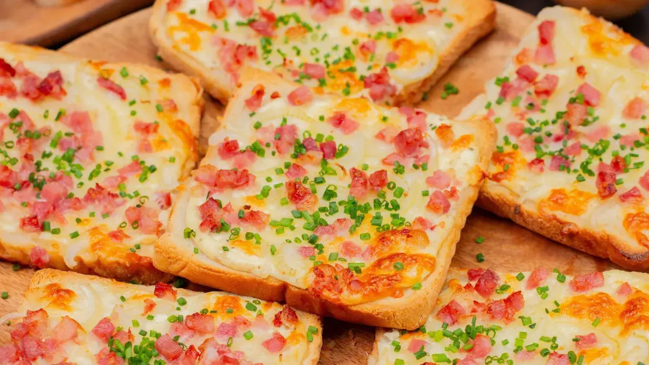 Mehrere Flammkuchen-Toast-Scheiben mit Speck, Zwiebeln und Schnittlauch belegt, auf einem Holzbrett.