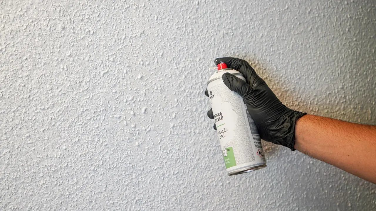 Mano con guante negro aplica spray para reparar gotelé. La pared tiene textura rugosa y blanca.