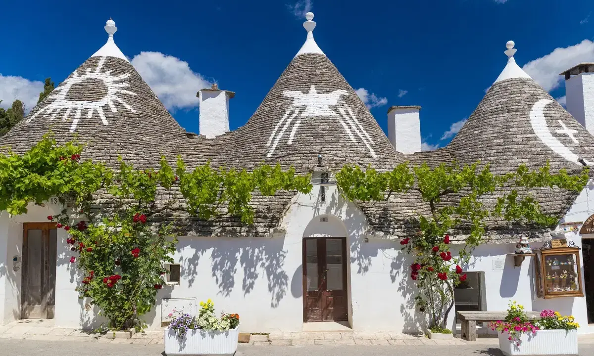 Trulli con simboli sui tetti, circondati da fiori e verde. Un'idea per scoprire **paesi da visitare vicino Alberobello**.