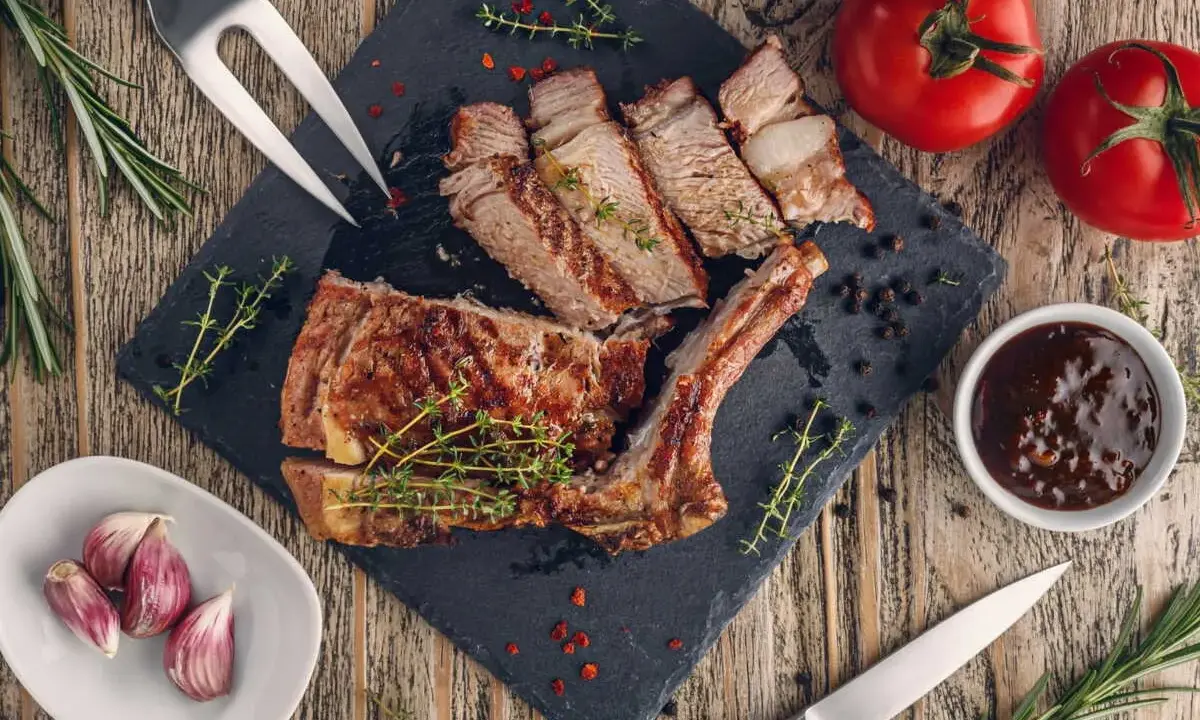 Côte de veau grillée tranchée sur une planche d'ardoise, garnie d'herbes, avec des tomates, de l'ail et une sauce.