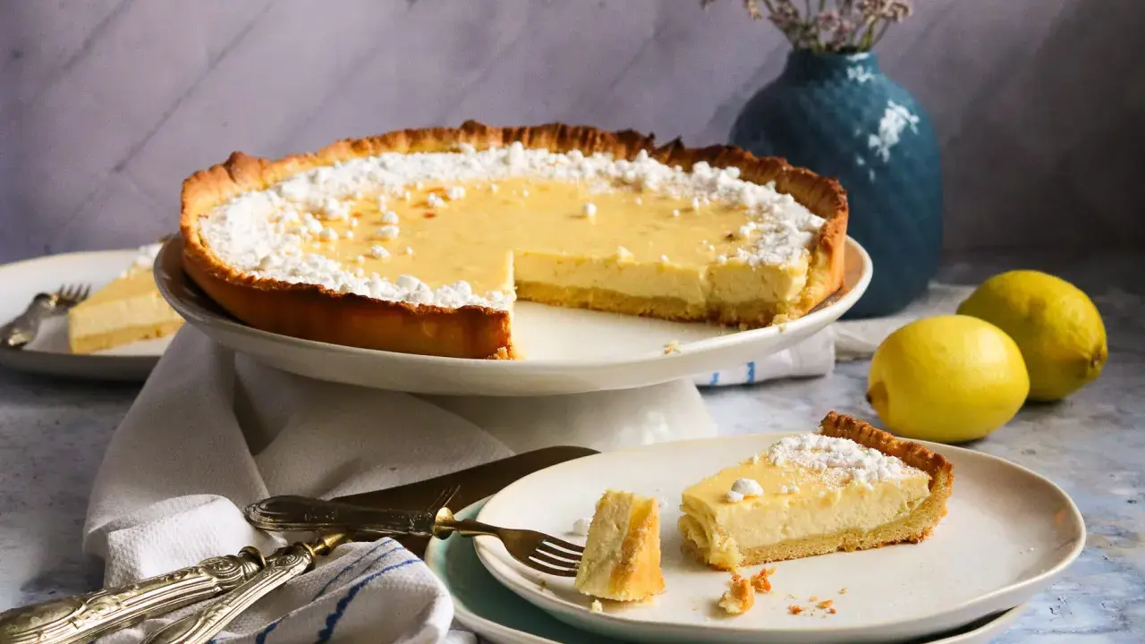 Ein köstliches Zitronentarte Rezept: Eine ganze Tarte und ein Stück auf einem Teller, bestäubt mit Puderzucker. Zwei Zitronen liegen daneben.
