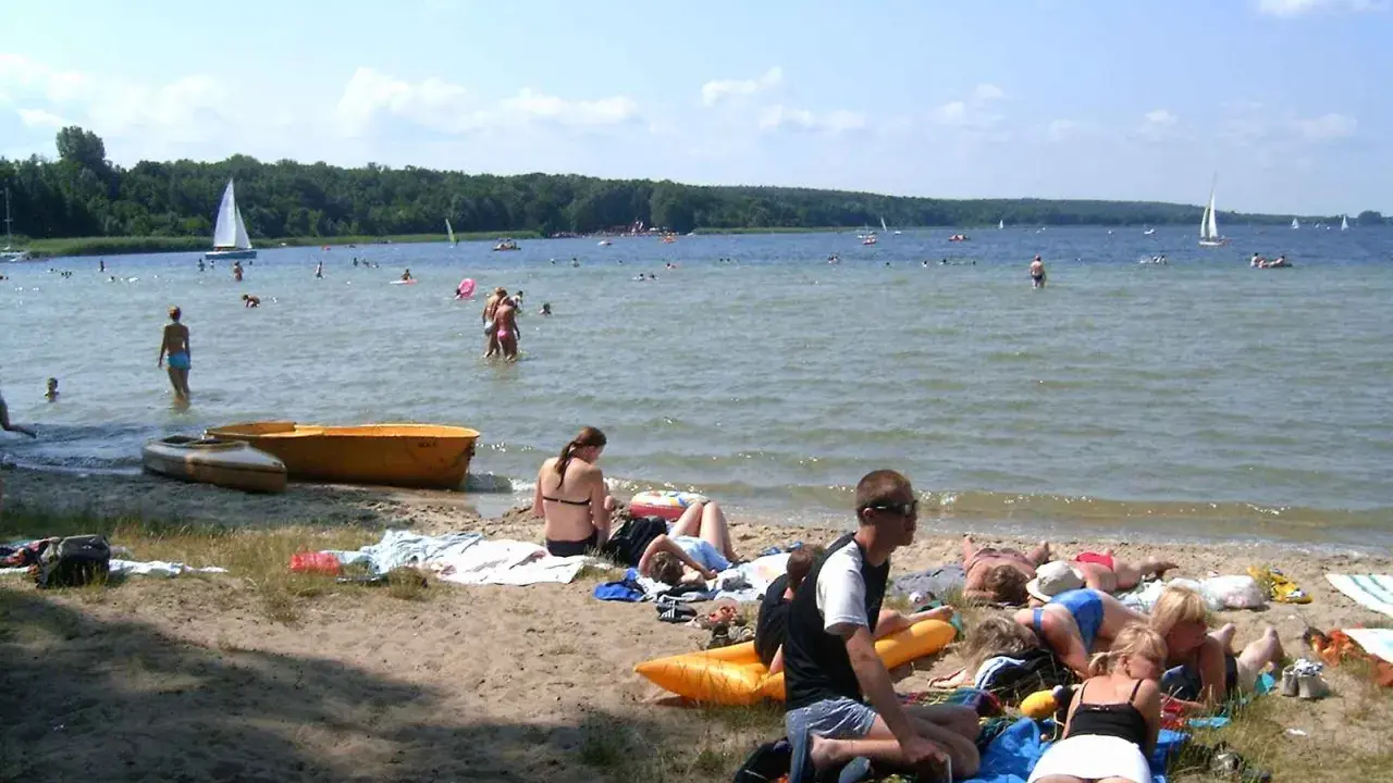 Ludzie odpoczywają na plaży i kąpią się w jeziorze. W tle żaglówki i las. To typowe boszkowo atrakcje w letni dzień.