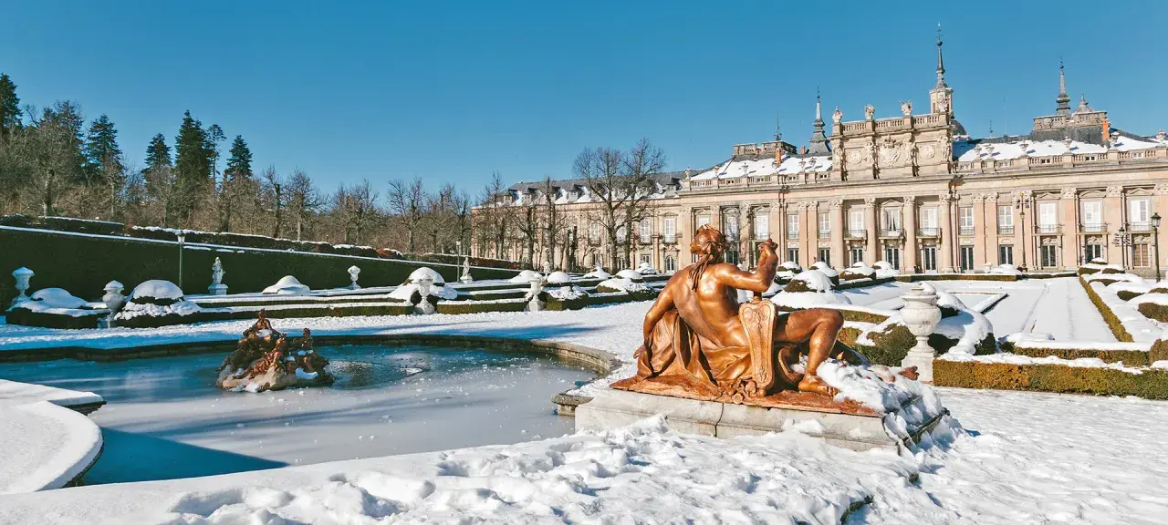 Schneebedeckte Gärten und ein gefrorener Brunnen vor einem Palast in Spanien im Winter. Eine bronzene Statue blickt auf die verschneite Landschaft.