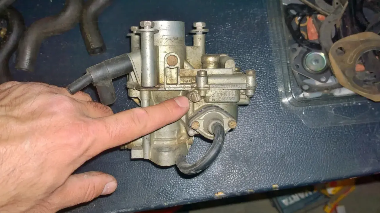 Main d'homme pointant un carburateur Zenith, possible cause d'un moteur 4 temps qui s'étouffe à l'accélération. Pièces de rechange visibles.