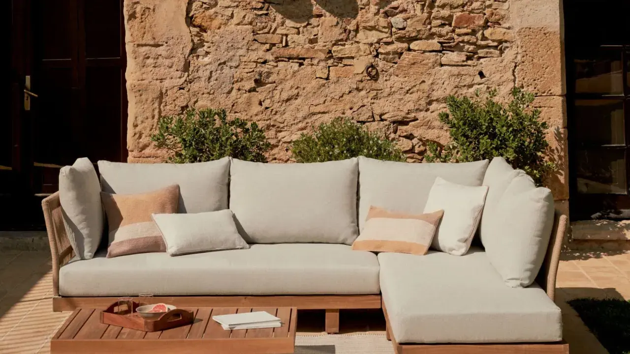 Sofá modular de exterior con cojines en tonos neutros, sobre una mesa de madera. La **decoración estilo natural** se realza con la pared de piedra y la vegetación.