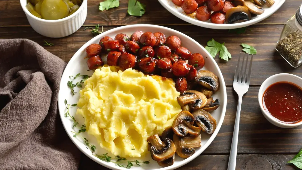 Talerz z puree ziemniaczanym, parówkami i pieczarkami, obok ogórki i sos.