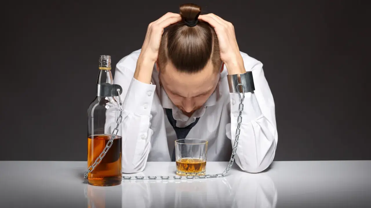 Mężczyzna w kajdanach, przykuty do butelki i szklanki z alkoholem, symbolizuje uzależnienie od piwa i jego niszczycielską siłę.