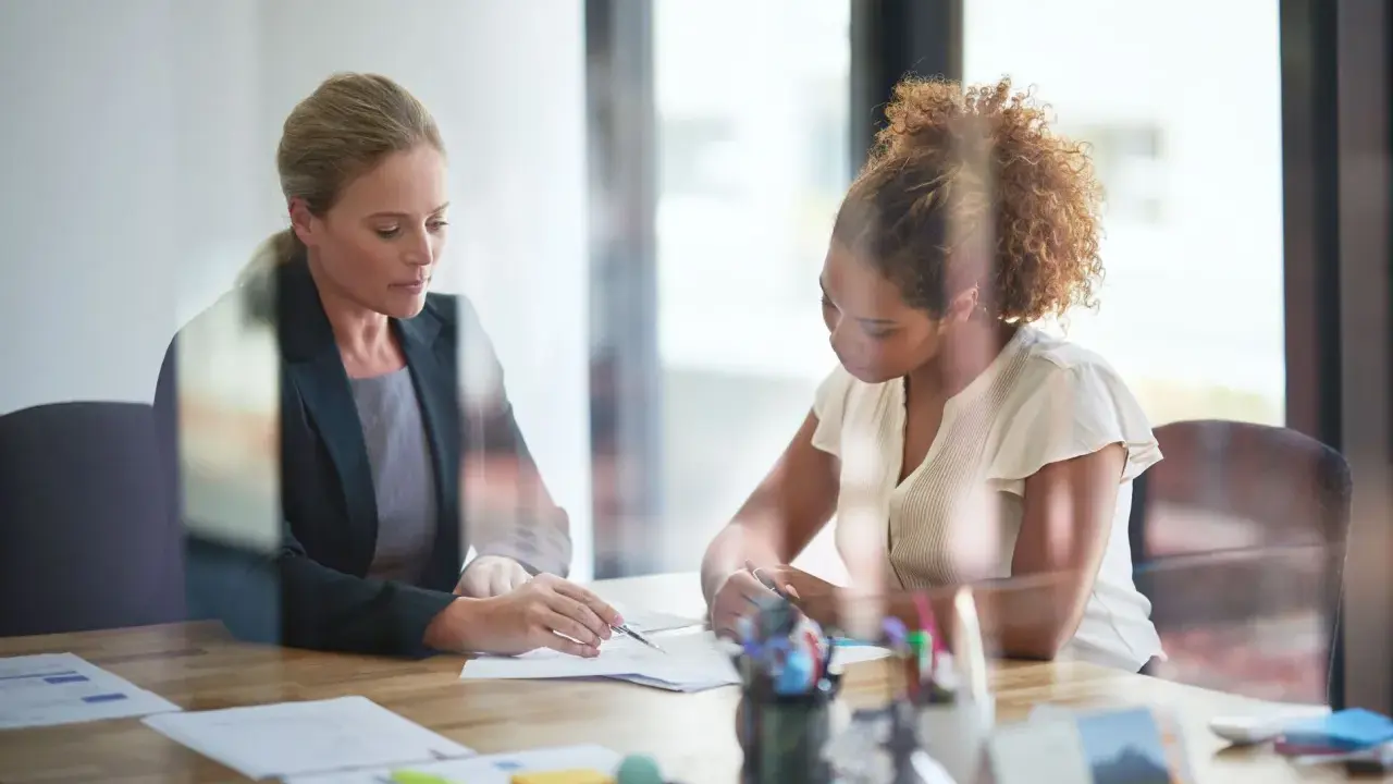 Deux femmes discutent de documents lors d'une réunion. L'une, blonde, porte un blazer sombre. L'autre, aux cheveux bouclés, est en chemisier clair. Leurs mains pointent vers les papiers, comme pour en comprendre le **signification**.