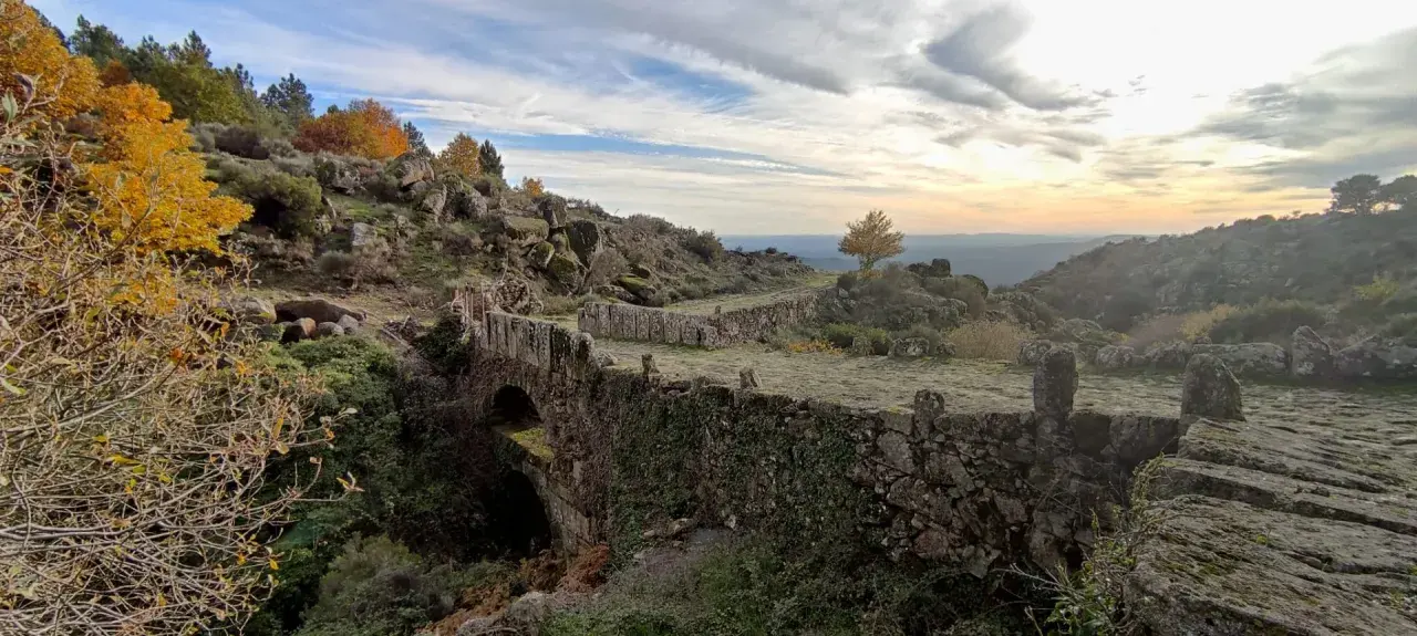 Herbstliche Farben in der Sierra de Gata. Ein alter Steinbogenbrücke, überwuchert von Efeu, führt durch eine felsige Landschaft.