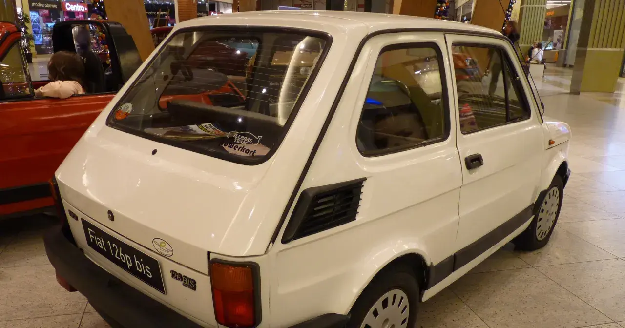 Biały Fiat 126p bis z naklejkami na tylnej szybie, stojący obok czerwonego samochodu.