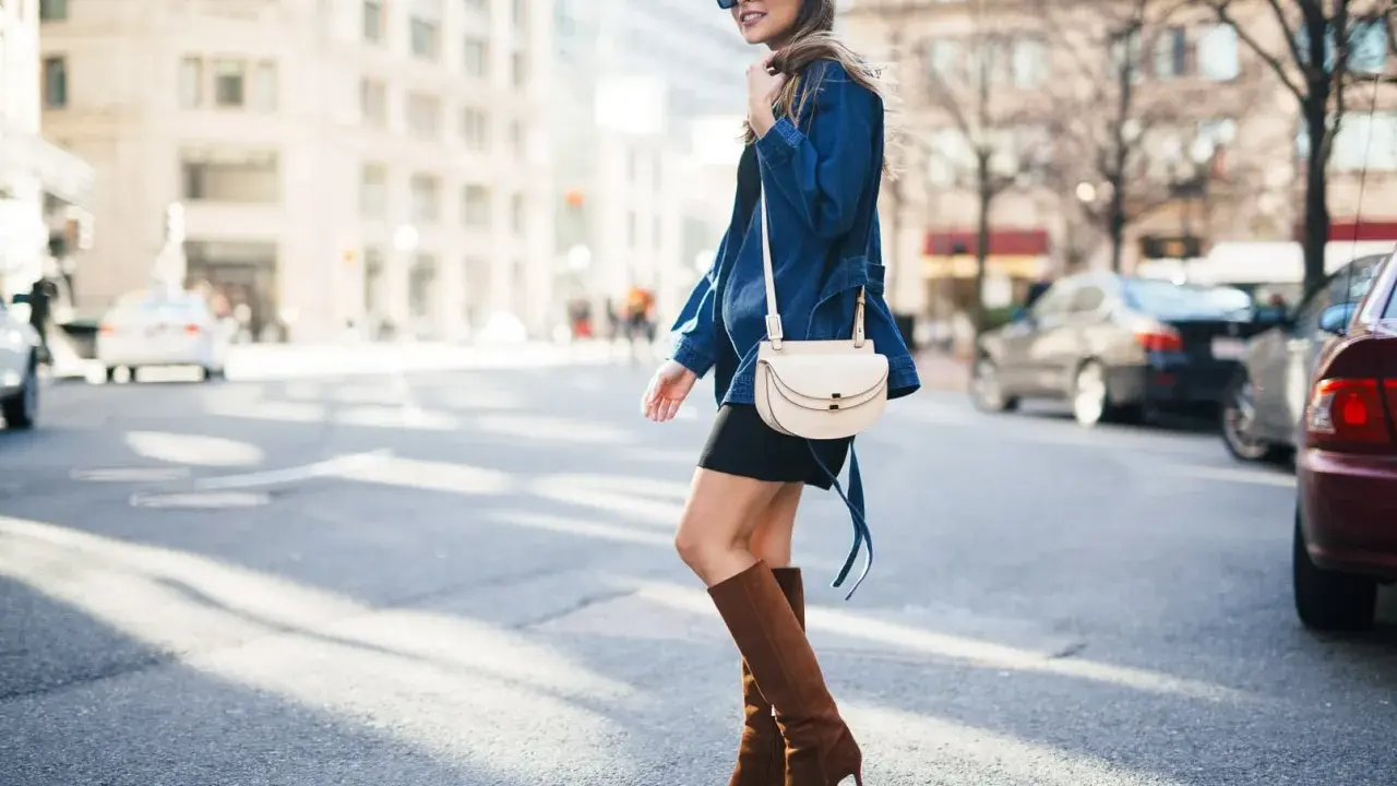A woman walks down a city street wearing a black dress, a denim jacket, and stylish brown boots.
