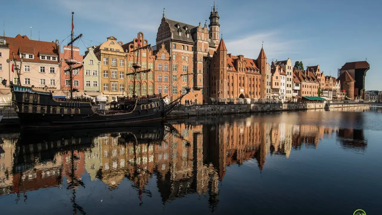 Gdańsk atrakcje: zabytkowy żaglowiec cumuje przy Motławie, w tle kolorowe kamienice i Żuraw. Odbicia budynków w wodzie.